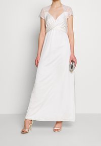 Robe longue blanche avec manches courtes en dentelle, détail noué à la taille, tissu lisse et drapé sur le côté. Associée à des talons argentés à brides et une pochette.