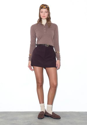 Žena nosící hnědý top s dlouhým rukávem, tmavou minisukni s opaskem, béžové ponožky, hnědé mokasíny a leopardí šátek na hlavě stojí na bílém pozadí.