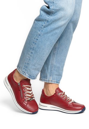 Voeten met rode leren sneakers met witte veters en zijritsen, gecombineerd met lichtblauwe spijkerbroek die net boven de enkels is afgeknipt.