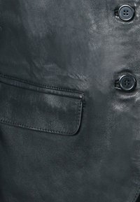 Giacca in pelle nera con una texture liscia, tasca frontale e due chiusure a bottone. Design strutturato con dettagli di cucitura discreti.