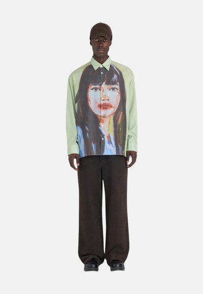 Chemise col polo en coton vert clair avec un design de portrait peint. Associée à un pantalon ample marron foncé et des chaussures noires.