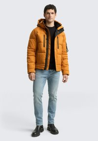 Geplüschte orange Jacke mit Kapuze, schwarzen Reißverschlussakzenten und vorderem Knopfverschluss. Kombiniert mit hellblauen Jeans und schwarzen Stiefeln.