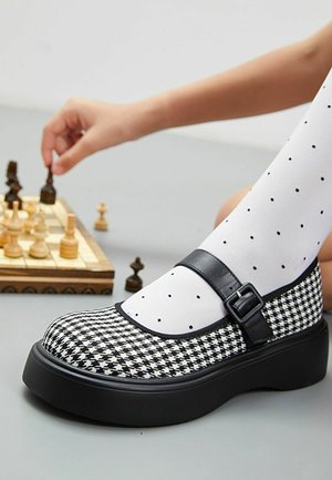 Zapato de patrón de pizarra en blanco y negro con punta redonda, correa de cuero y hebilla, y suela gruesa negra, combinado con medias blancas con lunares.