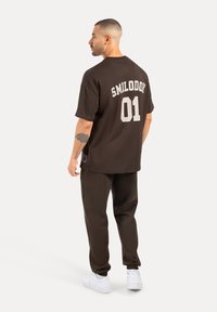 Camiseta de algodón marrón con "SMILODON 01" impreso en blanco en la parte posterior, combinada con pantalones oscuros y zapatillas blancas. Manga corta, corte relajado.