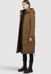 Barna puffernadrág kapucnival, oldalzsebekkel és cipzáros előlappal. Fekete részletek és textúra kontrasztok fokozzák a dizájnt. Fekete öltözék felett viselve.