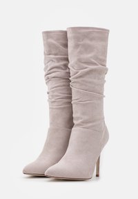 Par de botas altas de ante beige claro con punteras puntiagudas, cañas caídas y altos tacones de aguja sobre un fondo blanco.