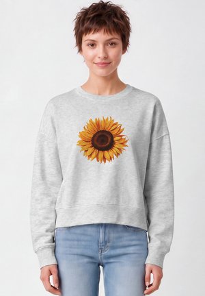 Jeune personne aux cheveux courts et bruns portant un sweat-shirt gris clair avec un grand motif de tournesol et un jean bleu clair.