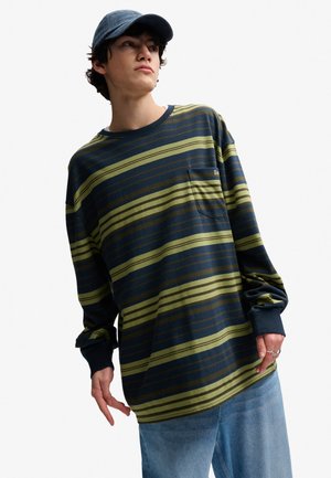 Ung person iført en blå og grønstribet langærmet trøje, lyseblå jeans, flere ringe og en mørkeblå baseballkasket, der kigger sidelengs.