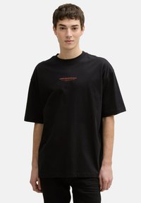 Black oversized t-shirt in cotton with a crew neck. Features red branding text on the chest. Short sleeves and a relaxed fit.
