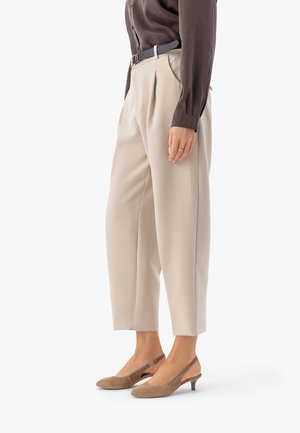 Person trägt beigefarbene, hoch taillierte Plisseehosen, ein dunkelbraunes Langarmshirt, das in die Hose gesteckt ist, einen schwarzen Gürtel und taupefarbene Wildleder-Kitten-Heel-Slingback-Schuhe.