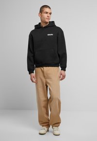 Schwarzer Kapuzenpullover mit einer Fronttasche, kombiniert mit beigen lässigen Hosen und weißen Sneakers. Einfaches Design ohne Muster.