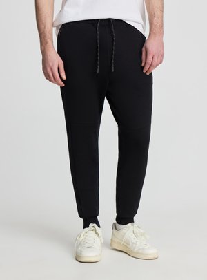 Mężczyzna ubrany w czarne spodnie dresowe ze sznurkiem w pasie i białe trampki, stojący na jasnym, jednolitym tle.