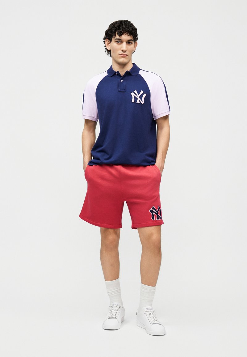 Jonge man die een marineblauw-witte New York Yankees polo draagt, rode korte broek met logo, witte sokken en witte sneakers, staand met handen in de zakken.