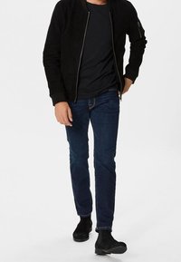 Chaqueta bomber de ante negro, camiseta negra, vaqueros azul oscuro y botines negros. La chaqueta tiene cierre frontal con cremallera y bolsillos laterales.