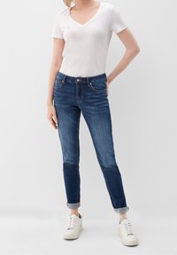 Camiseta ajustada de cuello en v, ligera y blanca, combinada con vaqueros azul oscuro ajustados con cintura a media altura y puños remangados, usada con zapatillas blancas.