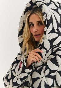 Nafukovaný černý a krémový kabát s velkými květinovými vzory, kapucí, prošívanou strukturou a těsným střihem, nošený s rukama blízko límce.