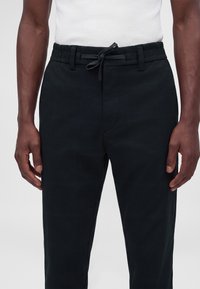 Homme portant un pantalon noir à cordon et un débardeur blanc côtelé sur un fond uni.
