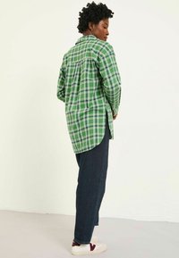 Green plaid flannel shirt with long sleeves, curved hem, and side slits, paired with dark denim jeans and white sneakers with maroon accents.