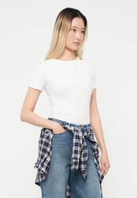 Fehér, rövid ujjú felső, kerek nyakkivágással, kék farmernadrággal és derékon megkötött kockás flanelinggel párosítva.