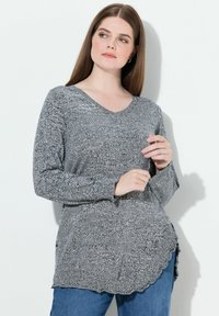 Suéter de punto gris con escote en V, mangas largas y dobladillo con forma de ola. Presenta una tela texturizada con un patrón de hilos mezclados. Combinado con jeans azules.