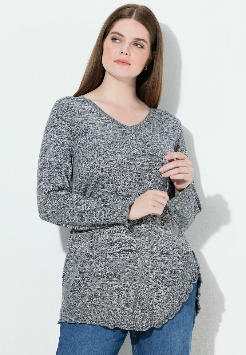 Suéter de punto gris con escote en V, mangas largas y dobladillo con forma de ola. Presenta una tela texturizada con un patrón de hilos mezclados. Combinado con jeans azules.