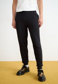 Sorte sweatpants med elastisk talje, sidelommer og et lille logo-patch, parret med sorte og beige mønstrede sko.