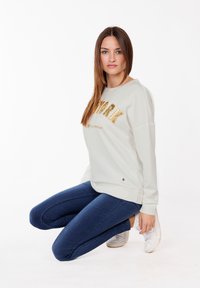 Sudadera gris claro con texto dorado, corte relajado, puños acanalados, combinada con vaqueros ajustados azul oscuro y zapatillas blancas.