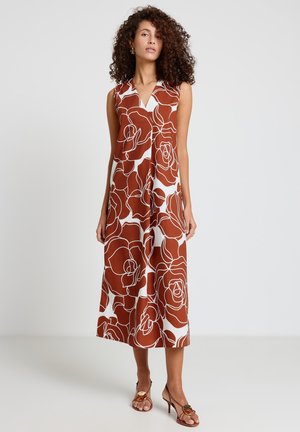 Donna con capelli ricci che indossa un vestito midi floreale marrone e bianco senza maniche e sandali marroni con tacco, in piedi davanti a uno sfondo semplice.