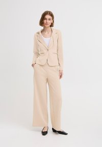 Béžový blazer s kapucňou a gombíkovým uzáverom, skombinovaný s voľnými béžovými nohavicami. Čierne špicaté baleríny dopĺňajú celý outfit.