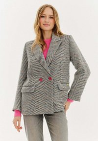 Žena s dlouhými blond vlasy, oblečená v šedém kostkovaném dvouřadém saku s červenými knoflíky přes jasně růžový svetr a šedé džíny.