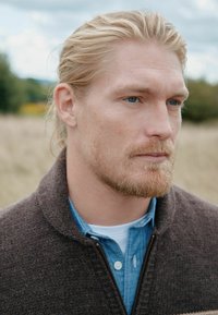 Mand iført en mørkebrun uldtrøje med høj krave over en lyseblå denimskjorte. Har lynlås og knapper, med struktureret stof.