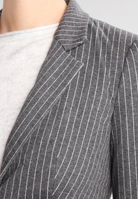 Blazer rayé gris à fines rayures blanches, avec un col structuré et un tissu texturé, partiellement visible sur un haut gris clair.