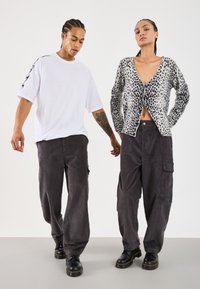 Two people stand side by side wearing black cargo pants and black shoes; one wears a white oversized T-shirt, the other a patterned cardigan.