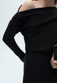Crni top bez ramena od glatkog materijala s dugim rukavima, s drobnim detaljima i uskim siluetom, uparen s crnom clutch torbom.