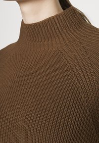 Närbild av en person som bär en brun ribbad stickad tröja med hög krage, som visar textur och detaljer vid axelsömmarna.