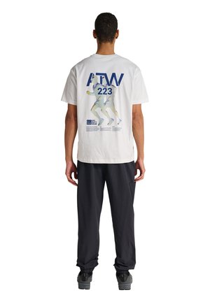 Mand iført hvid T-shirt med blå løbere og tekst "ATW 223" på ryggen, iført sorte bukser og grå sneakers, stående med ryggen mod os.
