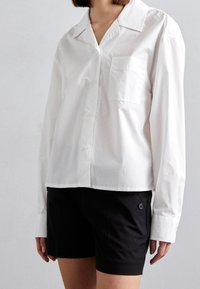 Camicia bianca con colletto e taschino sul petto, abbinata a pantaloni corti neri con bottoni laterali. Texture liscia, vestibilità comoda.