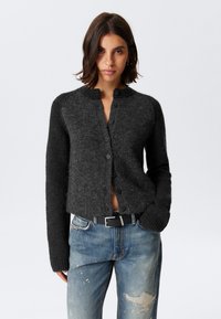 Donkergrijze cardigan met een ronde halslijn, voorknopen en geribde manchetten, gecombineerd met lichtgekleurde distressed jeans en een zwarte riem.