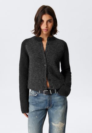Cardigan grigio scuro con scollatura rotonda, bottoni frontali e polsini a coste, abbinato a jeans chiari con strappi e una cintura nera.