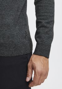 Suéter gris oscuro hecho de una tela de punto texturizada, con puños y dobladillo acanalados. Pequeña etiqueta de la marca visible cerca de la parte inferior en el lateral.