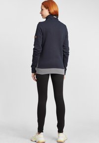 Pull en polaire bleu marine avec un col montant, un ourlet gris et des accents sur les manches ; associé à des leggings noirs et des chaussures de sport légères.