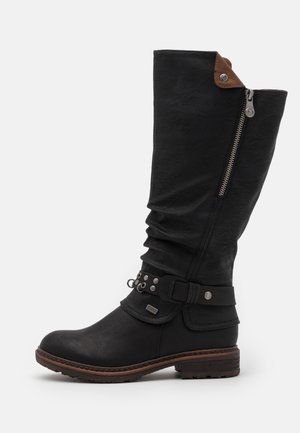 Botte en cuir noire mi-mollet avec fermeture éclair latérale, bordure marron en haut, sangle décorative avec clous et talon bas empilé sur fond blanc.