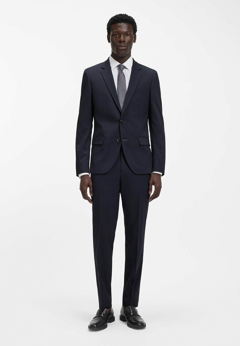 Completo a due pezzi blu navy con vestibilità su misura, giacca monopetto con revers a lancia, pantaloni a piega piana, abbinato a una camicia chiara e una cravatta fantasia.