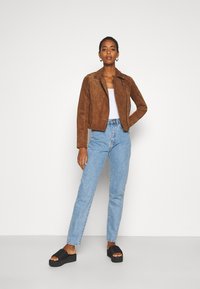 Jaqueta de camurça castanha com um design ajustado, jeans de perna reta azul claro e sandálias de plataforma pretas, contra um fundo neutro.