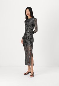 Robe en dentelle noire avec des motifs floraux en argent délicats, manches longues, silhouette ajustée et fente sur le côté, associée à des sandales noires à lanières.
