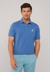 Koszulka polo w jasnoniebieskiej bawełnie z klasycznym kołnierzykiem, zapięciem na trzy guziki i małym haftowanym logo na lewej piersi. Krótkie rękawy.