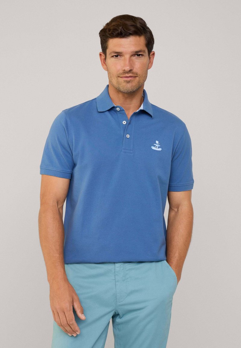 Koszulka polo w jasnoniebieskiej bawełnie z klasycznym kołnierzykiem, zapięciem na trzy guziki i małym haftowanym logo na lewej piersi. Krótkie rękawy.
