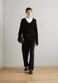 Suéter negro de punto con cuello en V sobre una camisa blanca de cuello, combinado con pantalones negros y zapatos negros brillantes con acentos dorados.