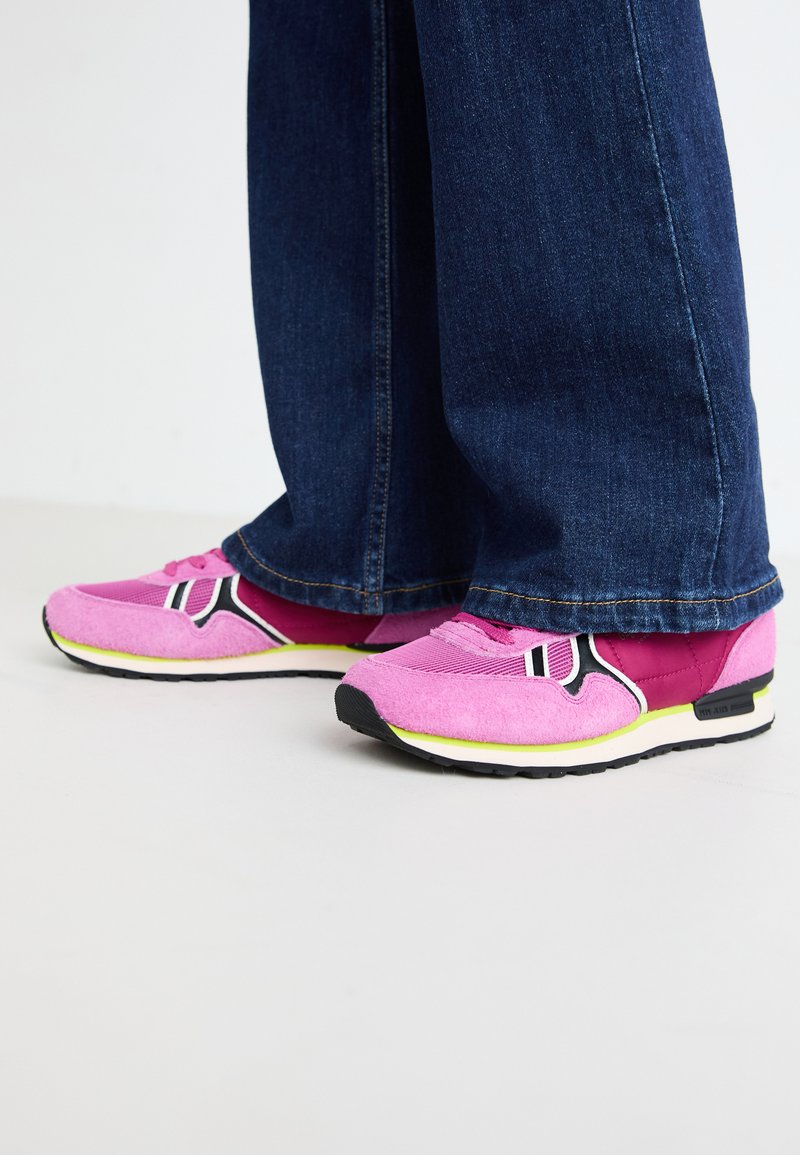 Sportliche Schuhe in Pink aus Wildleder und Mesh-Materialien, mit schwarzen und weißen Akzenten und einer gelben Sohle. Kombiniert mit dunkelblauen Jeans.