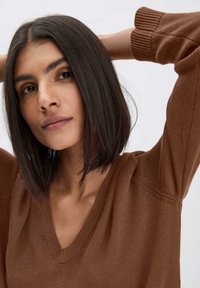 Brauner gestrickter Pullover mit V-Ausschnitt, strukturierten Ärmeln und lässiger Passform, getragen von einem Model mit glattem, schulterlangen Haar.